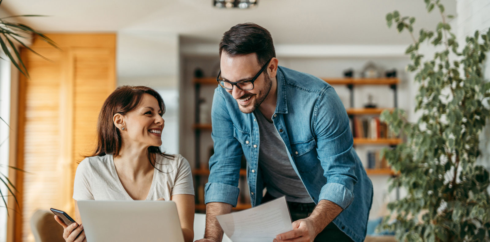 A man and a woman smiling and looking at a piece of paper and a laptop.